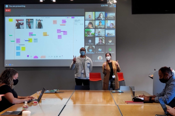 Me presenting in a Google conference room, masked, in front of a projected FigJam board covered in sticky notes, with a grid of remote attendees on screen beside it.