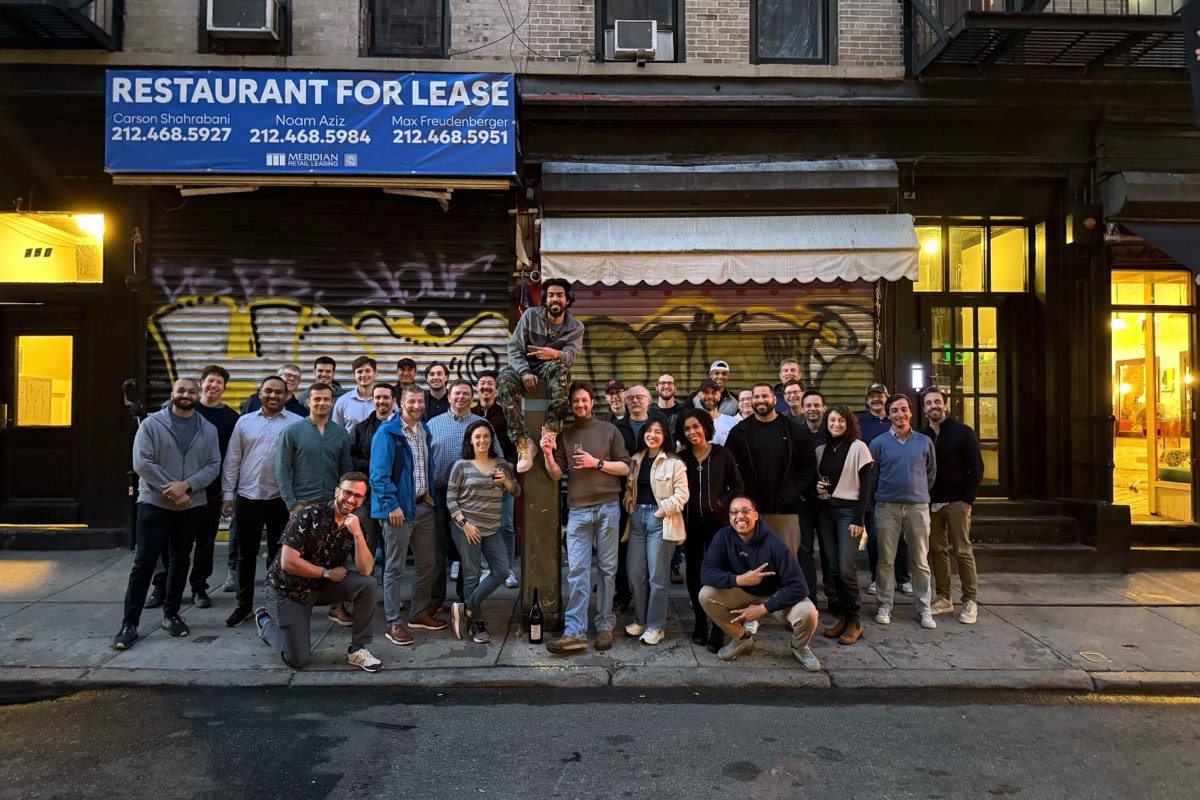 Most of the Runwise team crowded onto a New York sidewalk at dusk, in front of a graffitied shutter and a Restaurant For Lease banner, one person perched on a post above the group.